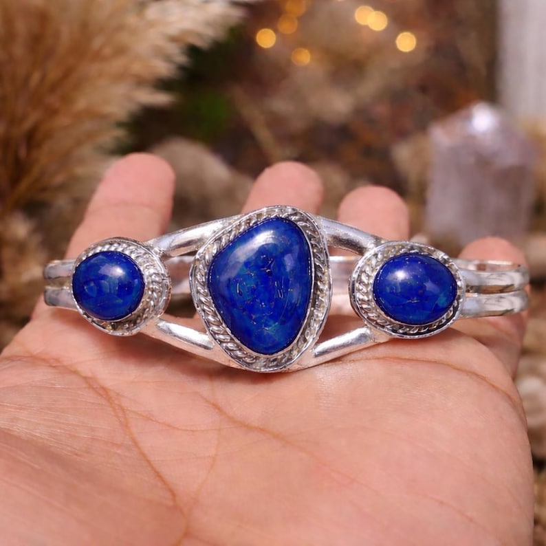 May include: A silver cuff bracelet featuring three vibrant blue lapis lazuli stones. The central stone is an irregular oval shape, while the flanking stones are round. The bracelet has a braided rope design around the stones.