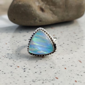 May include: A sterling silver ring with a triangular opal gemstone. The opal has iridescent blue and green hues. The ring's bezel is adorned with small, beaded details. The ring is set against a speckled surface with a blurred rock in the background.