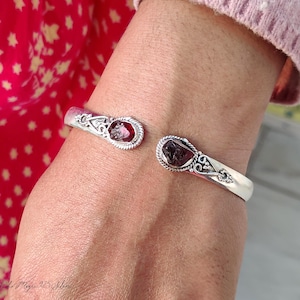 May include: A silver cuff bracelet featuring two dark red gemstones. The bracelet has an open design with decorative detailing around the stones. The gemstones are oval-shaped and set in a bezel. The bracelet is on a wrist.