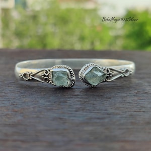Puede incluir: Un brazalete de plata con dos acentos de piedras preciosas verde claro. El brazalete est&aacute; abierto y tiene un dise&ntilde;o delicado.
