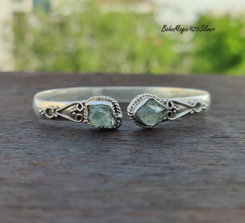 Puede incluir: Un brazalete de plata con dos acentos de piedras preciosas verde claro. El brazalete est&aacute; abierto y tiene un dise&ntilde;o delicado.