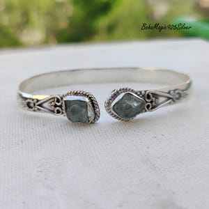Puede incluir: Un brazalete de plata con dos piedras de aguamarina azul claro engastadas en el centro. El brazalete tiene un dise&ntilde;o delicado y ornamentado.