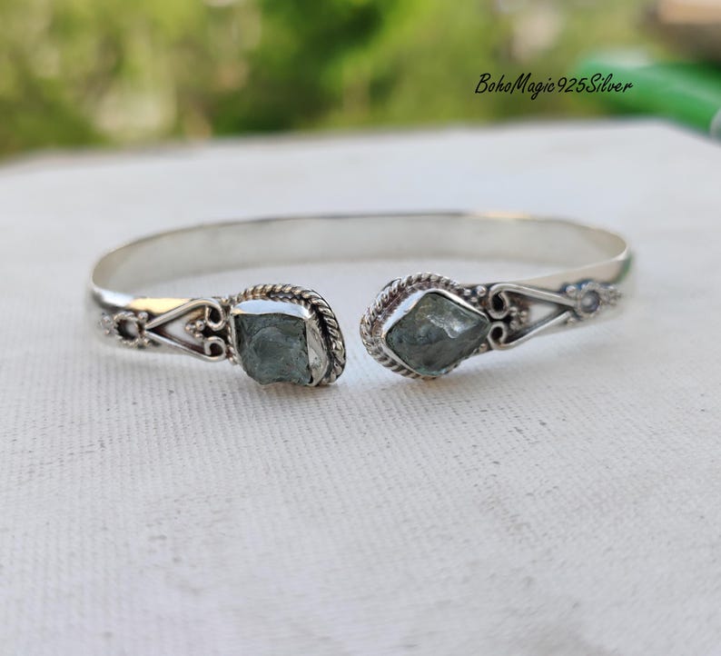 Puede incluir: Un brazalete de plata con dos piedras de aguamarina azul claro engastadas en el centro. El brazalete tiene un dise&ntilde;o delicado y ornamentado.