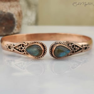 May include: A copper-colored open cuff bracelet with ornate detailing. The bracelet features two teardrop-shaped, blue-green gemstones at the ends. The ends of the bracelet have decorative filigree patterns. The text "Boho Magic 925 Silver" is in the upper right corner.