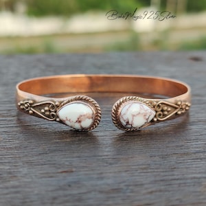 May include: Copper cuff bracelet with white and brown teardrop-shaped stones. The bracelet has decorative detailing near the stones and a simple, open design. The text "Boho Magic 925 Silver" is visible in the background.