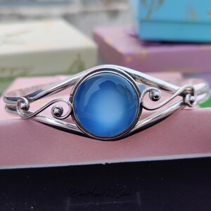 May include: A silver bangle bracelet with a large, round, light blue gemstone set in the center. The bracelet has a delicate, ornate design.