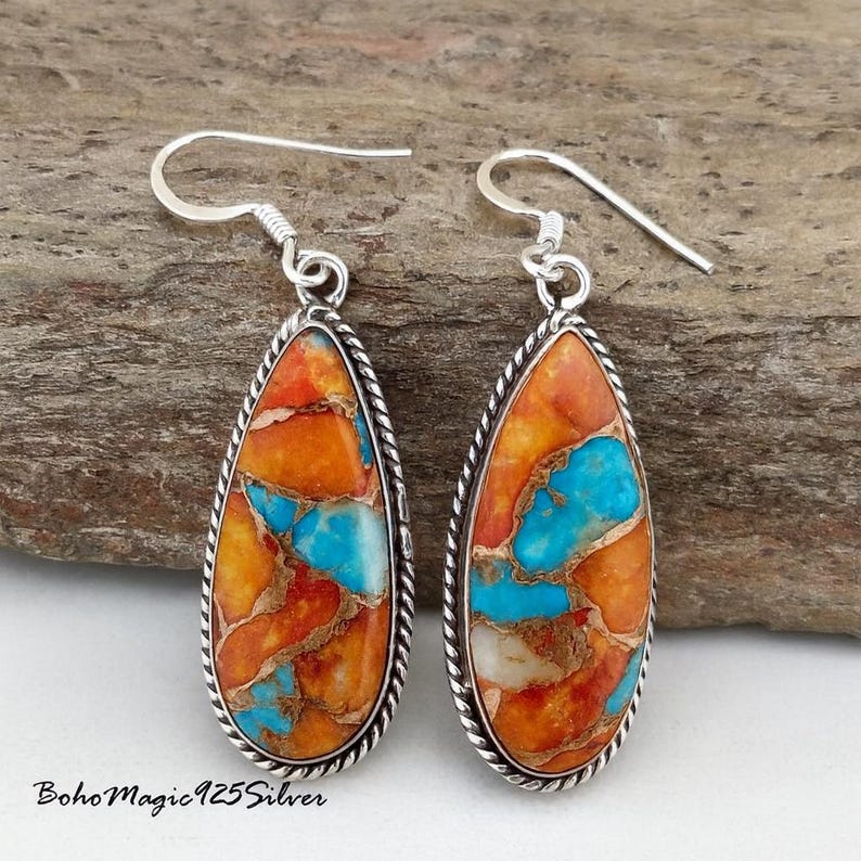 May include: Teardrop-shaped earrings with a silver frame and a colorful stone inlay. The stone features a mosaic of orange, turquoise, and white hues. The earrings have a hook closure. The text "Boho Magic 925 Silver" is visible at the bottom.
