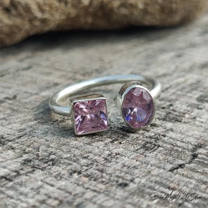 May include: A silver ring with two pink gemstones. One gemstone is square-cut, and the other is oval-shaped. The ring is set against a textured, gray surface. The ring is a piece of jewelry.