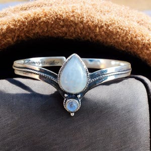 May include: A silver bracelet with a teardrop-shaped moonstone and a smaller round moonstone. The bracelet is designed in a V-shape and has a delicate, intricate design.