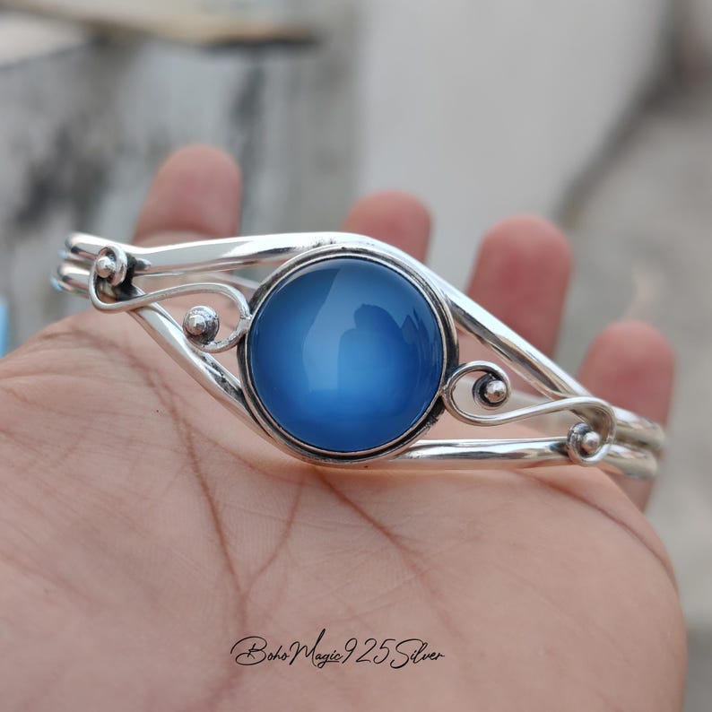 May include: A silver bangle bracelet with a blue cabochon gemstone. The bracelet has a delicate, ornate design with swirls and curves.