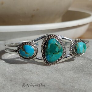 May include: A silver cuff bracelet featuring three turquoise stones. The bracelet has an oval turquoise stone on each end and a larger, triangular turquoise stone in the center. The stones are set in a silver setting with a rope-like border.