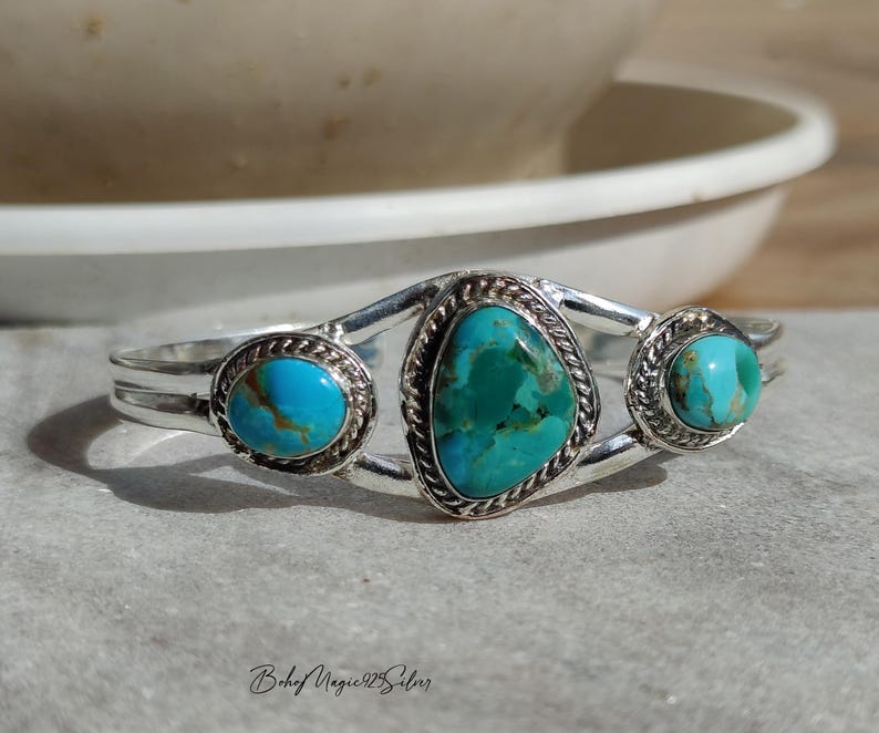 May include: A silver cuff bracelet featuring three turquoise stones. The bracelet has an oval turquoise stone on each end and a larger, triangular turquoise stone in the center. The stones are set in a silver setting with a rope-like border.