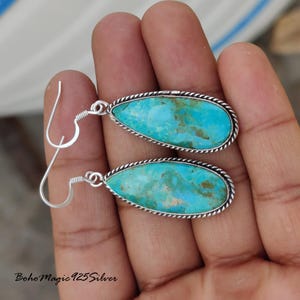May include: A pair of turquoise teardrop earrings. Each earring features a vibrant blue-green stone with brown veining, set in a silver-toned frame with a twisted rope design. The earrings have silver ear wires.