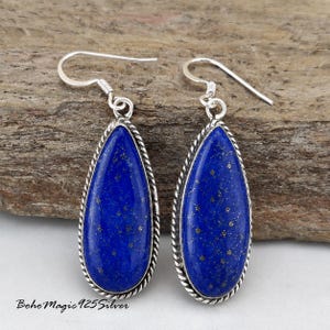 May include: A pair of teardrop-shaped earrings featuring deep blue lapis lazuli stones with silver-colored metal accents. The earrings have a rope-like border and a hook closure. The text "BohoMagic925Silver" is visible at the bottom.