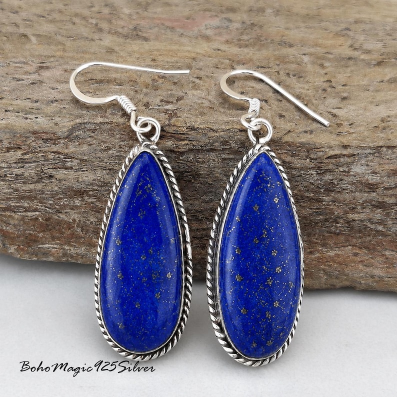 May include: A pair of teardrop-shaped earrings featuring deep blue lapis lazuli stones with silver-colored metal accents. The earrings have a rope-like border and a hook closure. The text "BohoMagic925Silver" is visible at the bottom.