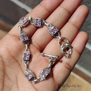 May include: A silver bracelet with square, pale purple gemstones set in decorative frames. The bracelet is linked with small connectors and has a toggle clasp. The gemstones are evenly spaced along the bracelet's length.