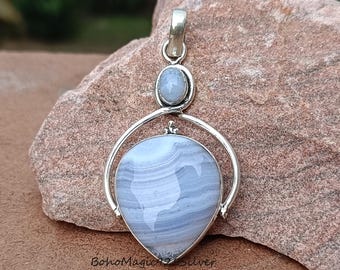 May include: A silver pendant featuring a blue agate stone. The stone is teardrop shaped and has a swirling pattern. The pendant is displayed on a rock background.