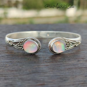 May include: A silver cuff bracelet with two oval opal stones. The bracelet has an open design with decorative filigree and rope-like detailing around the opals. The opals display a range of pastel colors.