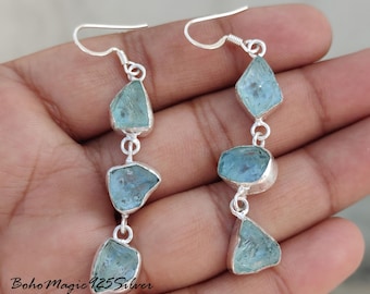 May include: Silver earrings with a dangling chain of four light blue aquamarine stones. The stones are irregular in shape and have a natural, raw look.