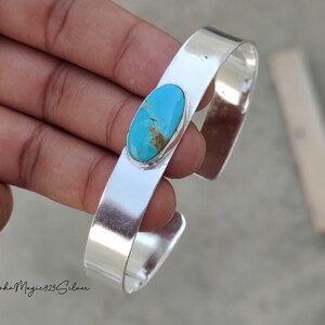 May include: A silver cuff bracelet featuring an oval turquoise stone. The turquoise stone has a blue and brown pattern. The bracelet is smooth and shiny. The text "BohoMagic925Silver" is at the bottom.