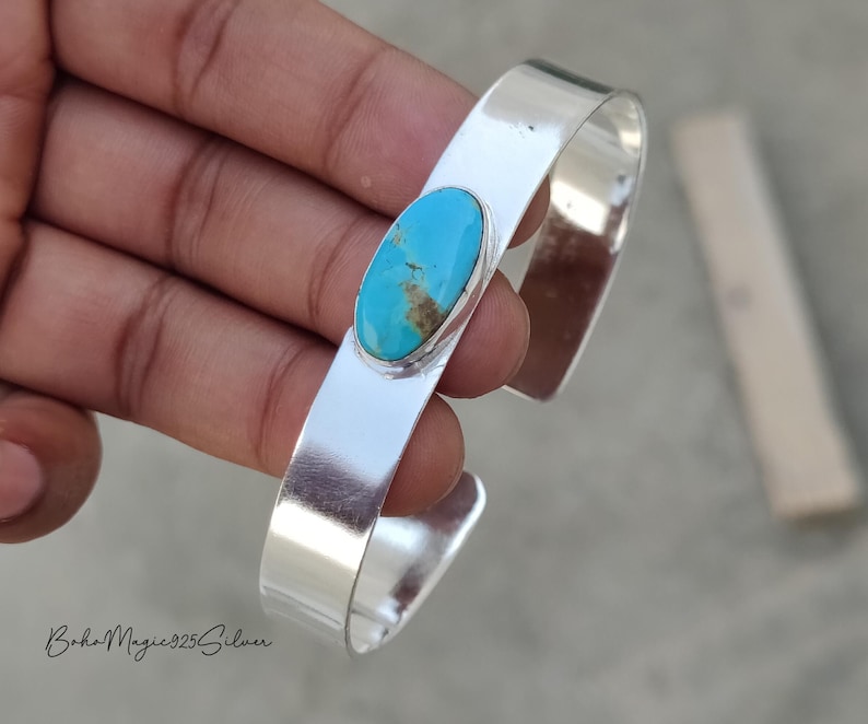 May include: A silver cuff bracelet featuring an oval turquoise stone. The turquoise stone has a blue and brown pattern. The bracelet is smooth and shiny. The text "BohoMagic925Silver" is at the bottom.