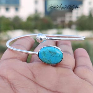 May include: A silver cuff bracelet with a large, oval turquoise stone. The open design has a smooth, polished finish and a rounded end opposite the gemstone. The turquoise stone has a natural, veined pattern.