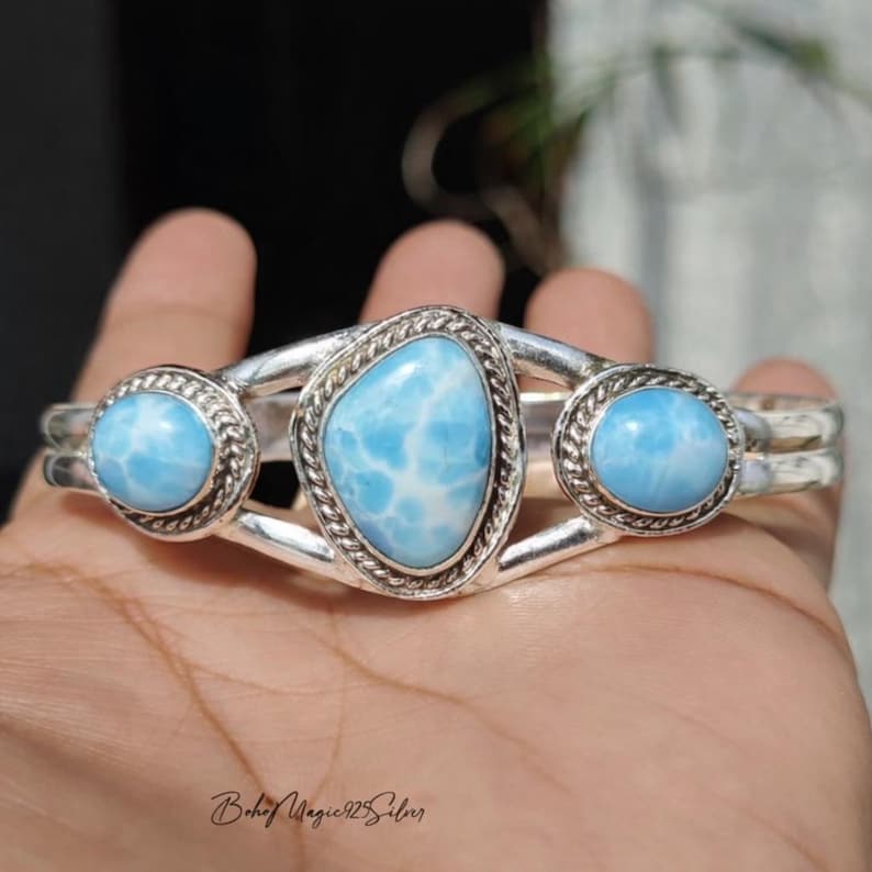 May include: A silver cuff bracelet featuring three light blue larimar stones. The central stone is an irregular oval shape, while the flanking stones are round. The bracelet has a polished finish and a delicate design.