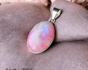 May include: A silver pendant featuring a large oval-shaped iridescent gemstone with pink, white, and blue hues. The pendant is on a soft, light purple fabric.