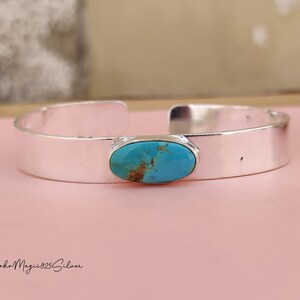 May include: A silver cuff bracelet featuring an oval turquoise stone. The turquoise stone has brown and blue tones. The bracelet is displayed on a pink surface. The text "BohoMagic925Silver" is at the bottom.