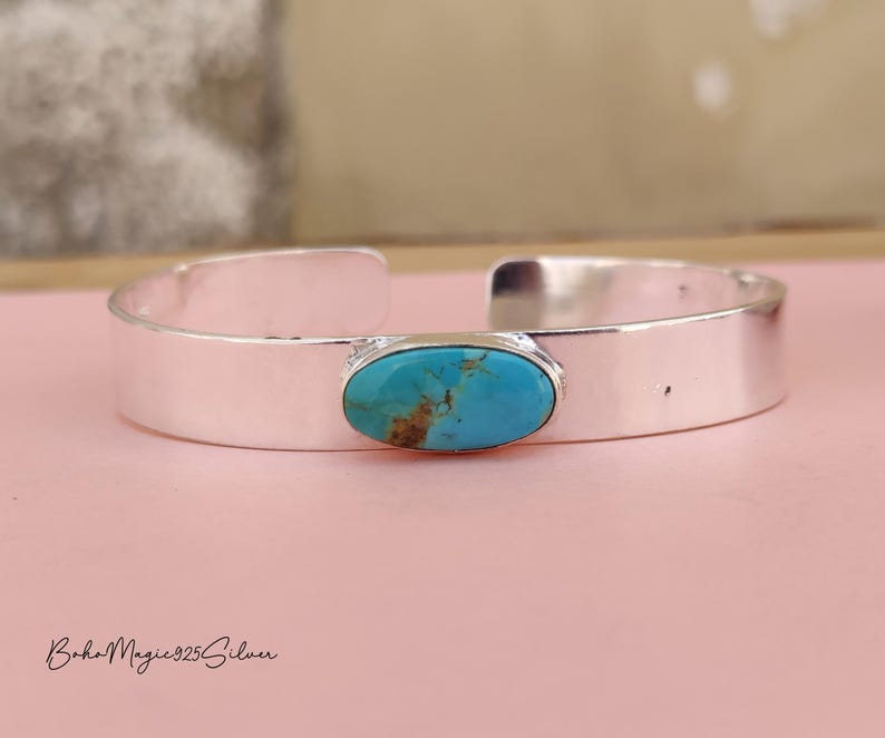 May include: A silver cuff bracelet featuring an oval turquoise stone. The turquoise stone has brown and blue tones. The bracelet is displayed on a pink surface. The text "BohoMagic925Silver" is at the bottom.