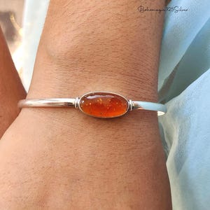 Peut inclure: Un bracelet jonc en argent avec une pierre précieuse ovale de couleur orange. La pierre précieuse a un aspect tacheté. Le bracelet est sur un poignet, avec un tissu bleu clair en arrière-plan.