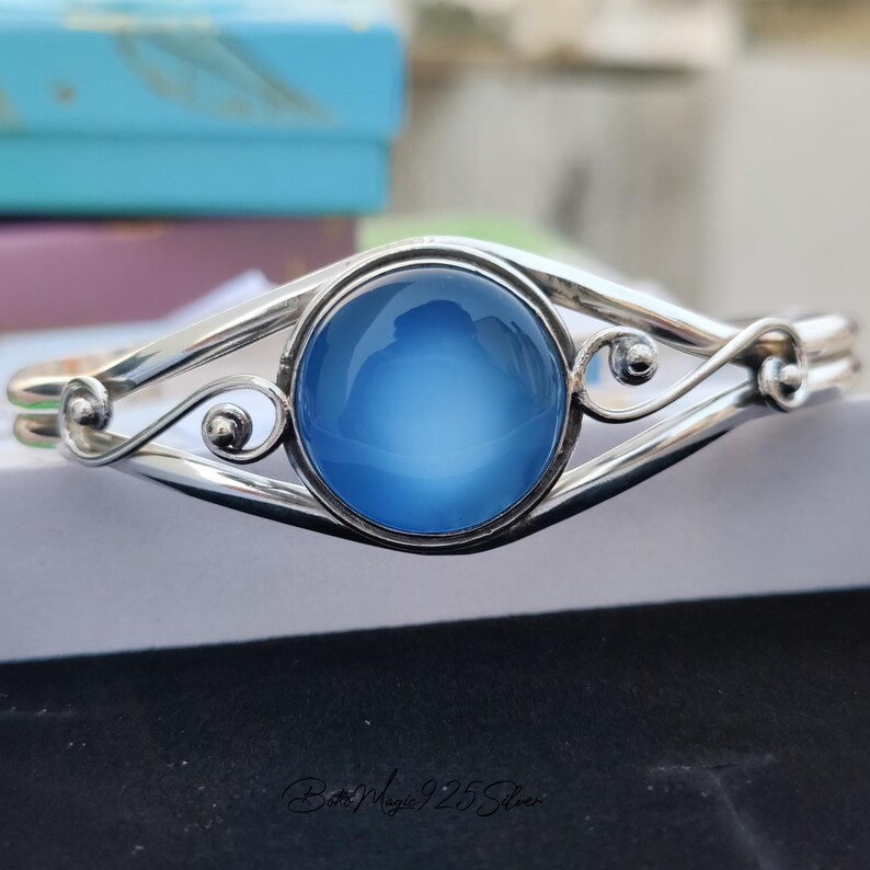 May include: A silver bangle bracelet with a blue cabochon gemstone. The bracelet has a delicate, ornate design.