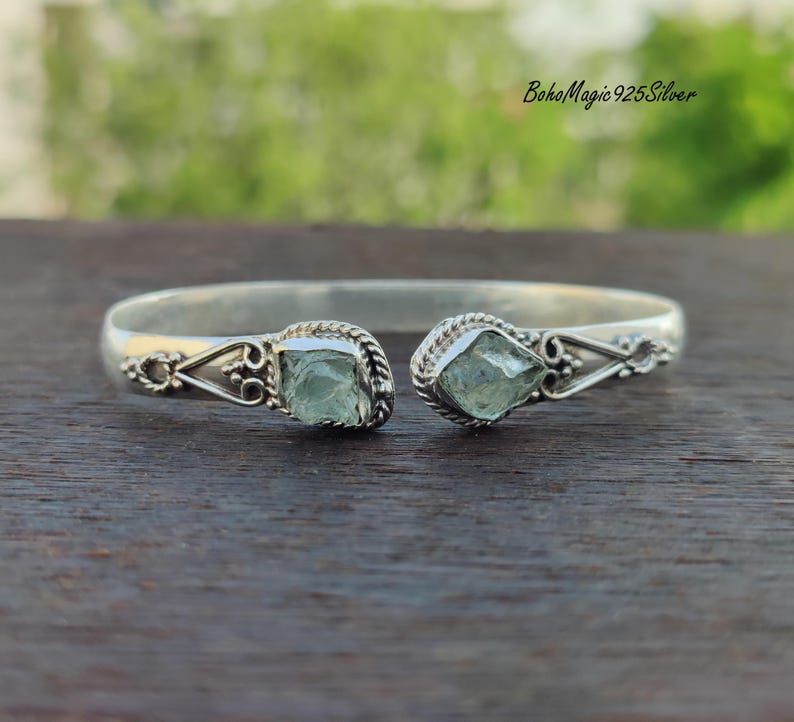 Puede incluir: Un brazalete de plata con dos piedras de aguamarina verde claro engastadas en los extremos. El brazalete est&aacute; abierto y tiene un dise&ntilde;o delicado.