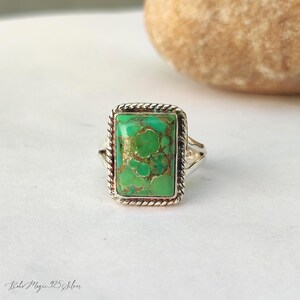 May include: A silver ring featuring a rectangular green stone with gold veining. The stone is set within a twisted rope-style border. The ring is displayed on a white surface. The ring is a piece of jewellery.