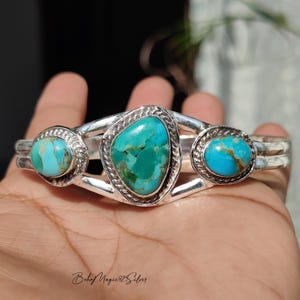 May include: A silver cuff bracelet featuring three turquoise stones. The central stone is a large, irregular shape, flanked by two smaller, round stones. The turquoise has a vibrant blue-green color, set in a silver band.