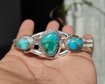 Pode incluir: Uma pulseira de punho de prata com três pedras turquesas. A pedra central tem uma forma irregular, ladeada por duas pedras redondas menores. A turquesa tem uma cor azul-esverdeada vibrante, definida em uma faixa de prata.