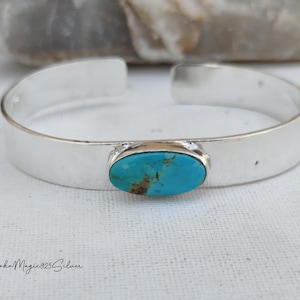 May include: A silver cuff bracelet featuring an oval turquoise stone. The stone is set in a silver bezel. The bracelet has a wide, minimalist design. The turquoise stone displays a striking blue-green hue. The bracelet is approximately 15 cm in circumference.