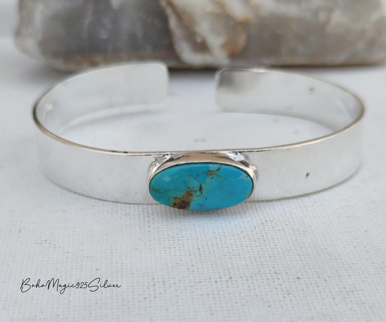 May include: A silver cuff bracelet featuring an oval turquoise stone. The stone is set in a silver bezel. The bracelet has a wide, minimalist design. The turquoise stone displays a striking blue-green hue. The bracelet is approximately 15 cm in circumference.