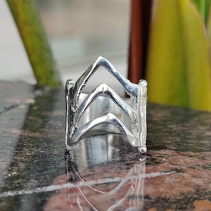May include: A close-up of a silver ring with a unique design. The ring features three chevron-shaped elements stacked on top of each other. The ring is made of a shiny, metallic material and is set against a blurred background.