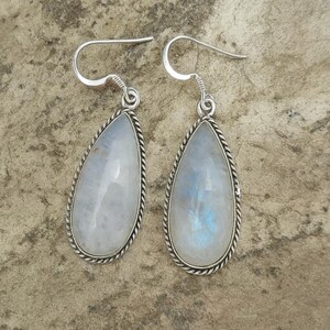 May include: A pair of teardrop-shaped earrings featuring iridescent moonstone gemstones. Each stone is set in a silver-toned metal frame with a twisted rope design around the edges. The earrings have a simple hook closure.