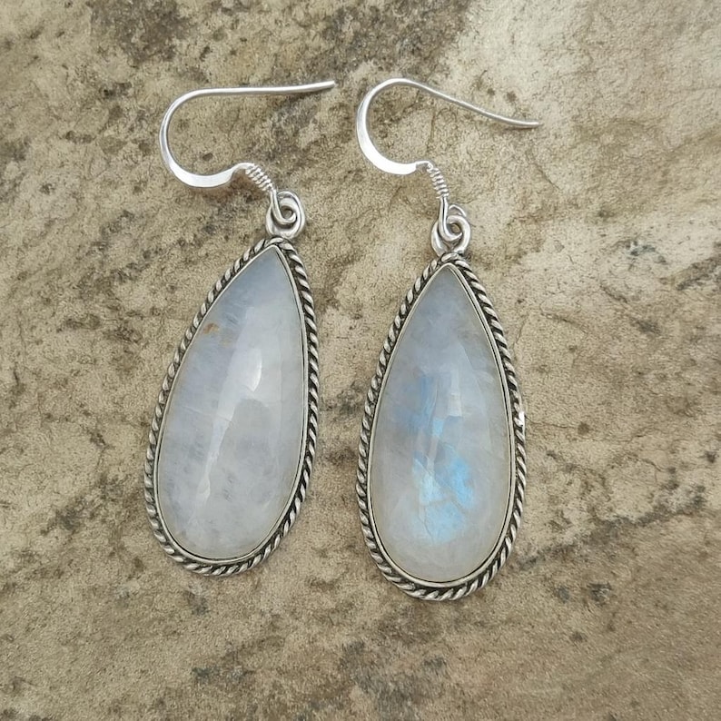 May include: A pair of teardrop-shaped earrings featuring iridescent moonstone gemstones. Each stone is set in a silver-toned metal frame with a twisted rope design around the edges. The earrings have a simple hook closure.