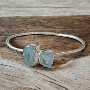 May include: A silver bangle bracelet with two aquamarine stones attached. The stones are raw and have a light blue colour.