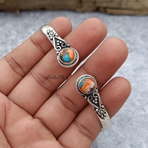 May include: Two silver bangle bracelets with ornate designs and a round turquoise and orange stone set in each. The bracelets are open and have a simple, elegant design.