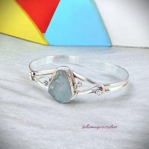 Peut inclure: Un bracelet jonc en argent avec une pierre d'aigue-marine bleu clair sertie dans un motif délicat et tourbillonnant. Le bracelet est ouvert et réglable.