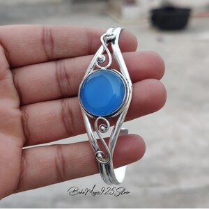 May include: A silver bangle bracelet with a blue gemstone set in a decorative silver frame. The bracelet is open and adjustable.