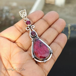 May include: A silver pendant with a large teardrop-shaped ruby gemstone and two smaller ruby gemstones. The pendant is set in a silver wire frame.