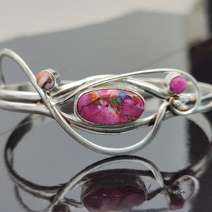May include: A silver bracelet with a pink and brown oval-shaped gemstone, a smaller pink gemstone, and a smaller white and orange gemstone. The bracelet has a delicate, flowing design.