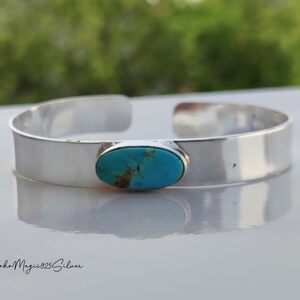 May include: A silver cuff bracelet featuring an oval turquoise gemstone. The bracelet has a polished finish and a minimalist design. The text "BohoMagic925Silver" is at the bottom. The bracelet is a stylish accessory.