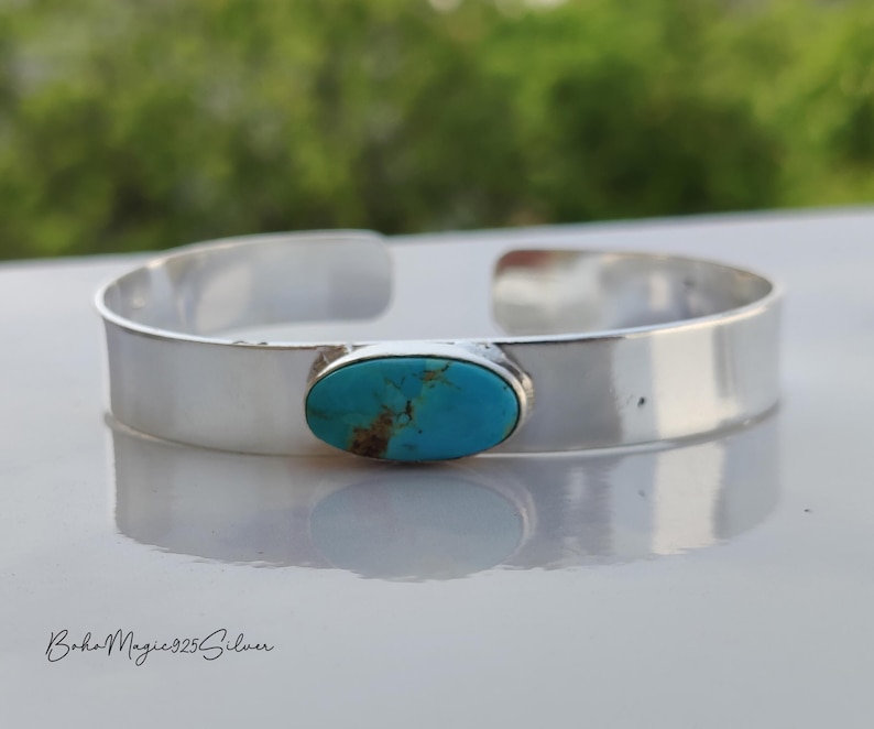 May include: A silver cuff bracelet featuring an oval turquoise gemstone. The bracelet has a polished finish and a minimalist design. The text "BohoMagic925Silver" is at the bottom. The bracelet is a stylish accessory.