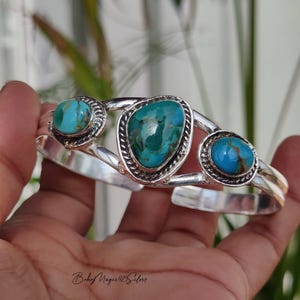 May include: A silver cuff bracelet featuring three turquoise stones. The stones are oval and teardrop-shaped, set in ornate silver bezels. The bracelet has a polished finish and a hinged design.